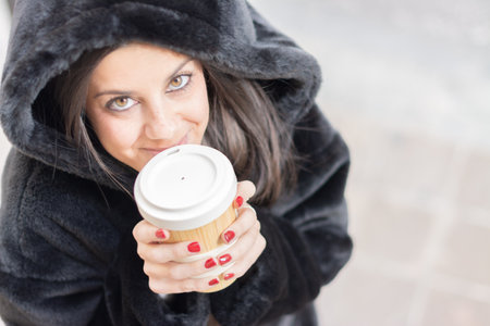 View from above of a girl looking at camera, on the street holding a reusable cup of coffee with both hands, feeling coldの写真素材