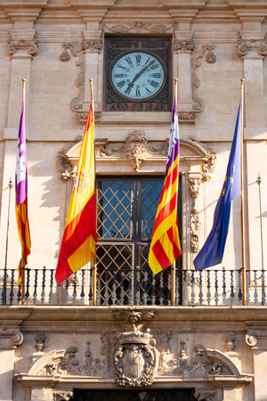 Palma, Mallorca, Balearic Islands, Spain. July 21, 2022 - Balcony on the facade of the town hall, with the flags of the European Community, the Spanish Community and the Balearic Community, with the clock and the stone coat of arms of the cityのeditorial素材