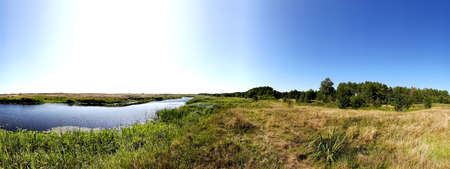Panorama of the river and meadows with herbs against the blue sky. Still green grass and river vegetation and withered dry grasses in the pasture.の写真素材