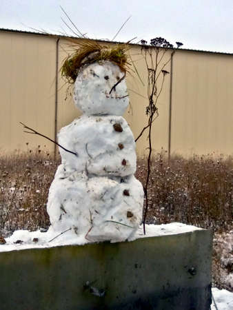 Unhappy snowman made of snow in winter. Dried herbs as decor. On a concrete foundation against the background of an industrial building and a sad sky.の写真素材