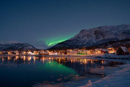 A small village nestled in a snowy valley is illuminated by the green glow of the Aurora Borealis. The reflection of the lights in the frozen water creates a stunning scene.の素材