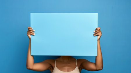 A person holds a large blue sign in front of their face, obscuring their facial features. The sign is blank and could be used for any message or text. The individual is wearing a light-colored tank top and their arms are extended, holding the sign securely. The background is a solid blue, creating a simple and clean visual.の素材