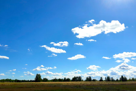 A clear blue sky filled with fluffy white clouds stretches over a peaceful countryside landscape, illuminated by sunlight.の写真素材