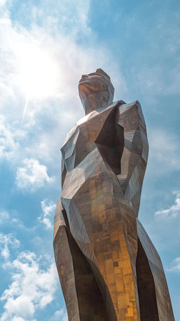 The towering abstract sculpture rises majestically against a vibrant blue sky, illuminated by sunlight. Surrounded by greenery, it captivates viewers with its striking form and artistic design.の素材
