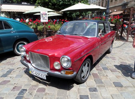 Lviv, Ukraine - June 28, 2014 A vintage red sedan VOLVO 162 DE LUXE sits elegantly on cobblestone streets in a bustling plaza. Nearby, colorful flowers and lush trees enhanceのeditorial素材