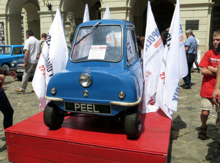 Lviv, Ukraine - June 28, 2014 A charming blue Peel P50 is showcased on a red platform at a bustling car show, surrounded by promotional flags and intrigued visitors admiring the unique vehicle's design and history.のeditorial素材