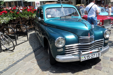 Lviv, Ukraine - June 28, 2014 A vintage GAZ-M20 Pobeda turquoise car is parked in a lively town square filled with colorful flowers and pedestrians. Sunlight brightens the sceneのeditorial素材