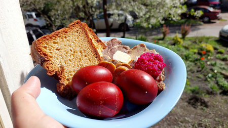 Colorful Easter plate held outdoors showcases red decorated eggs, slices of bread, meat, and a...