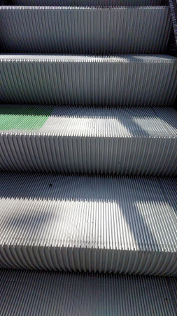 Escalator stairs ascend in a public area, sunlight casting shadows on the metallic surfaces. The scene captures the movement and functionality of urban transit.の写真素材