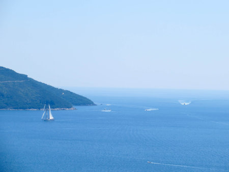A sailboat glides peacefully across the tranquil waters of the Bay of Kotor, surrounded by lush green hills under bright blue skies in Herceg Novi, Montenegro.の写真素材