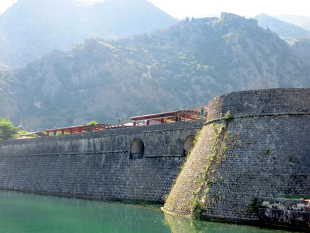 Visitors admire the ancient fortifications of Kotor, with stone walls reflecting in the clear waters of Boka Kotorska under the bright sun.の写真素材