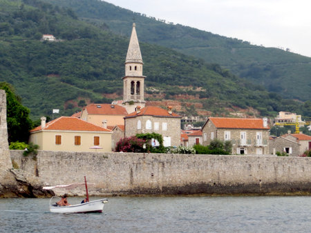 Visitors enjoy a serene day in Budva, Montenegro, as a small boat navigates along the coast beside the historical medieval town, showing its charming architecture and landscape.の写真素材