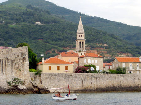 Budvas old town showcases stunning medieval architecture against a backdrop of lush hills. A small boat glides by, adding charm to this picturesque coastal landscape.の写真素材