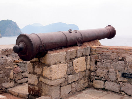 An old cannon stands on stone walls in Petrovac, Montenegro, providing a view of the calm bay and distant hills, evoking a sense of history and tranquility.の写真素材