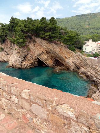 Clear blue waters of a rocky bay in Petrovac, Montenegro reveal stunning geological formations surrounded by lush greenery on a beautiful day.の写真素材