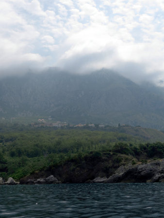 Dark cliffs rise from the calm waters of the Adriatic Sea near Montenegro, with a misty sky and lush greenery contrasting against the rocky shore.の写真素材