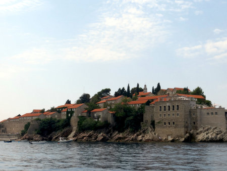 Sveti Stefan island resort in Montenegro showcases stunning red-tiled roofs amidst lush greenery, while boats gently float on calm waters under a clear sky.の写真素材