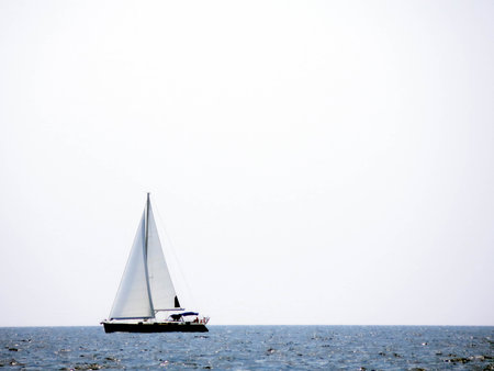 A sailing yacht with its sails raised navigates the tranquil sea on a sunny day. The wide expanse of water stretches under a clear sky, creating a serene atmosphere.の写真素材