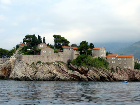 Explore the charming Sveti Stefan island resort in Montenegro, featuring stone buildings and lush greenery, set against a backdrop of serene waters on a cloudy day.の写真素材