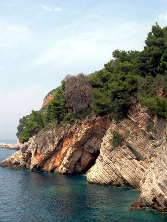 Glistening turquoise waters frame towering rocky cliffs in Petrovac, Montenegro, where lush greenery thrives along the rugged coastline under a clear sky.の写真素材