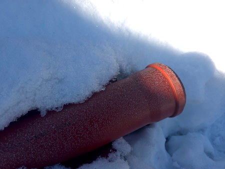 A red sewer pipe is partially buried under white snow. The scene shows winter conditions with...