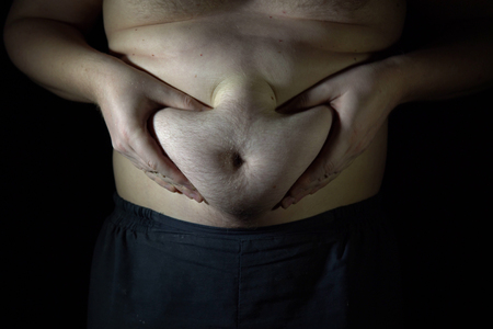 The fat man checks his big belly, he is obese and overweight. Close-up of a big male stomach on dark background.の写真素材