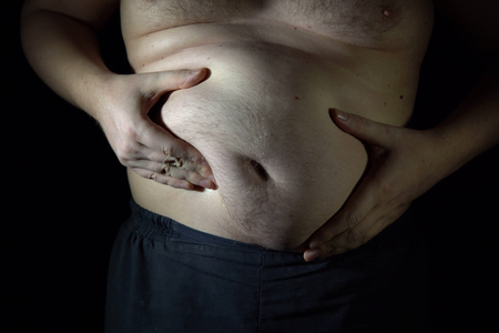 The fat man checks his big belly, he is obese and overweight. Close-up of a big male stomach on dark background.の写真素材