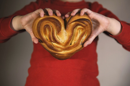 An unidentified child in a red t-shirt with long sleeves holds a sweet bun in the form of a heart.の写真素材