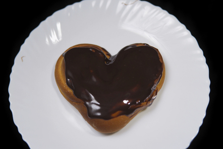 Top view of a heart shaped bun chocolate covered, baking lies on a white plate on a black background.の写真素材