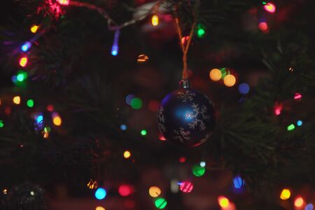 Blue xristmas ball with a silver snowflake weighs on branch of festive artificial Christmas tree among the colorful lights of garlands.の写真素材