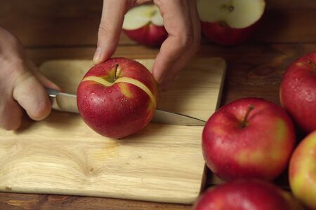 Hand with kitchen knife cuts ripe red apple on light wooden cutting board.の写真素材