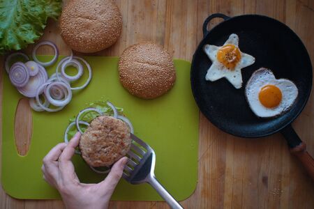 A top view of the process of cooking a hamburger at home, a man's hand puts a fried cutlet on half a bun, next to a star-shaped and heart-shaped fried eggs in the pan.の写真素材