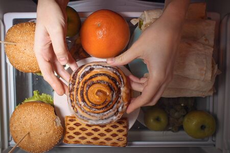 Top view of women's hands holding a sweet bun over the refrigerator shelf with burgers shawarma and fruit, flatlay.の写真素材