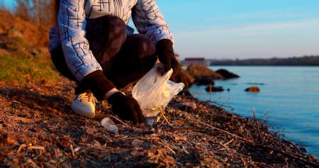 Young female volunteer collects garbage on the see shore in the evening at sunset in early spring or autumn, selective soft focusの写真素材