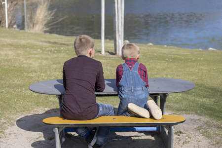Rear view of young boys with blond hair sitting on old beach picnic table facing ocean wearing plaid shirtの写真素材