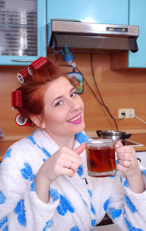 Young attractive red-haired woman with curlers drinking tea in the kitchenの写真素材