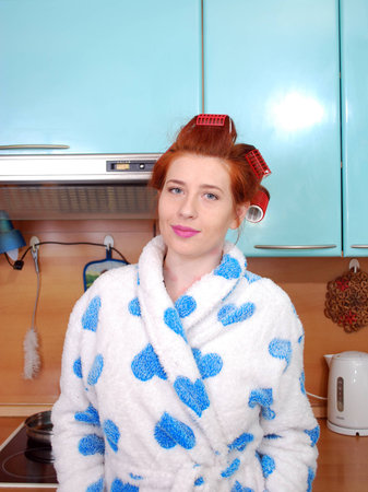 The young attractive girl with red hair in a dressing gown and with wound hair curlers costs in kitchen and smilesの写真素材