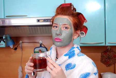 The attractive girl with red hair with a clay mask and hair curlers in hair costs in kitchen in a dressing gown and has hot teaの写真素材