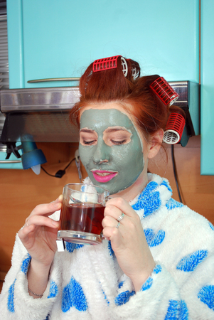 The attractive girl with red hair with a clay mask and hair curlers in hair costs in kitchen in a dressing gown and has hot teaの写真素材
