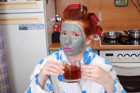 The attractive girl with red hair with a clay mask and hair curlers in hair costs in kitchen in a dressing gown and has hot teaの写真素材