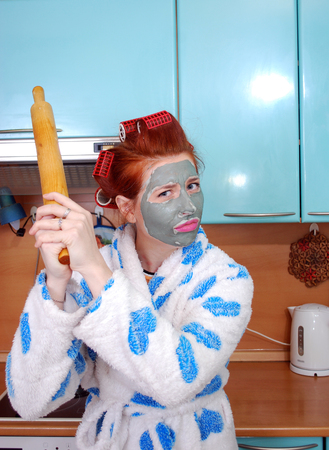 The young emotional housewife with red hair, in hair curlers, and with a clay mask on a face dissatisfied, threatens having threatened a rolling pinの写真素材