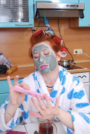 The young girl with red hair in hair curlers with a clay face pack in kitchen does manicureの写真素材