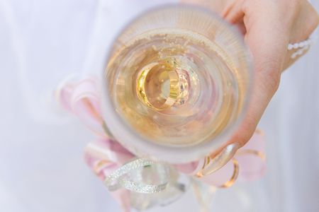 a hand of a bride holds a glass full of champagne and twwo gold rings in itの写真素材
