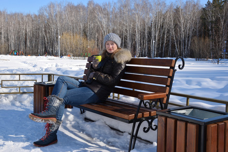 The girl sits on a bench in the park and drinks hot tea from a  bottle.の写真素材