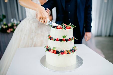 wedding ceremony. Hands of newlyweds cut a white three-tiered cake with strawberries and blackberriesの写真素材