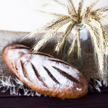 Rye bread lies on a wooden table covered with sacking next to a bunch of rye and wheat in a glass...