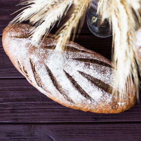 Rye bread lies on a wooden table covered with sacking next to a bunch of rye and wheat in a glass...