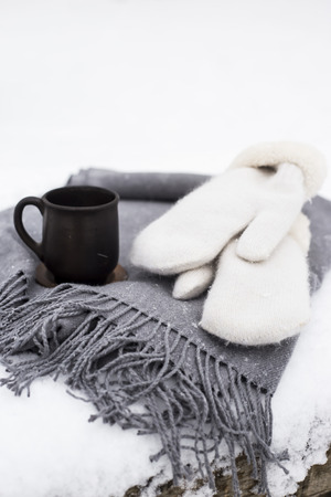 A cup of coffee on a gray plaid on a snowy table next to the white mittensの写真素材