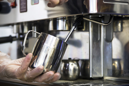 A barista at a cafe churns milk in a metal jug using a caper of  professional coffee machine. Coffee house concept. The process of making cappuccino or latteの写真素材