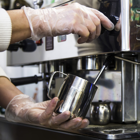 A barista at a cafe churns milk in a metal jug using a caper of  professional coffee machine. Coffee house concept. The process of making cappuccino or latteの写真素材